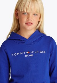 Szőke gyermek, kék Tommy Hilfiger kapucnis pulóvert visel, rajta a logó és az "EST. 1985" felirat, enyhén oldalra néz, fehér háttéren.