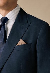 Marineblaues Sakko mit Reverskragen, strukturiertem Stoff und einem Einstecktuch im hellbeigen Muster. Das Hemd hat blau-weiß gestreifte Muster.