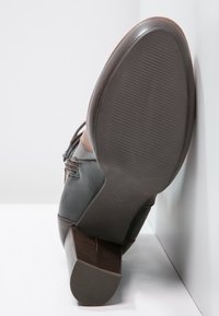 Bruine leren enkelboot met blokhak, gezien vanaf de zoolzijde, steunt tegen een witte muur en oppervlak.