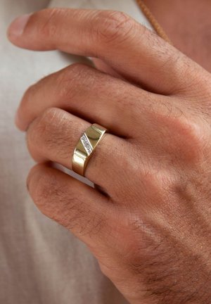 Bague en or avec un design ondulé et une ligne diagonale de petites pierres claires. La main est légèrement bronzée avec une texture visible.