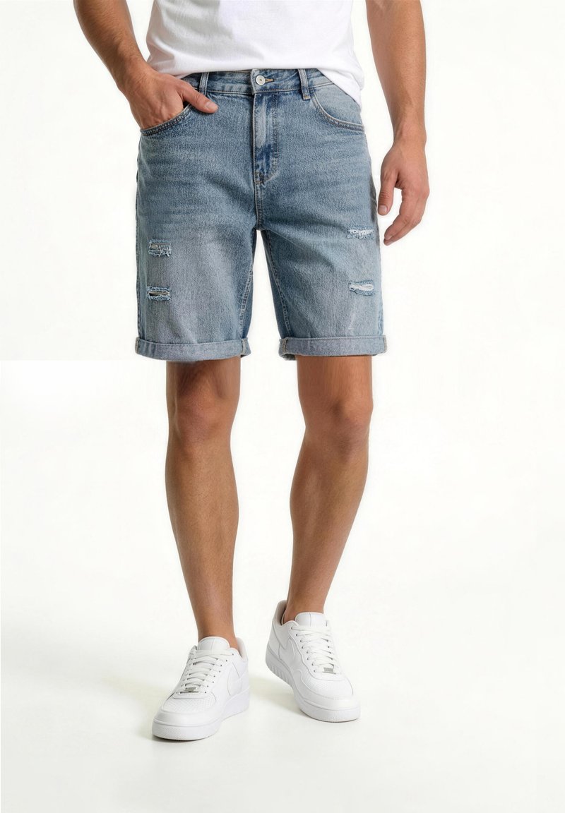 Homme portant un short en denim bleu clair retroussé avec des détails usés, des baskets blanches et un t-shirt blanc, debout avec une main dans la poche.