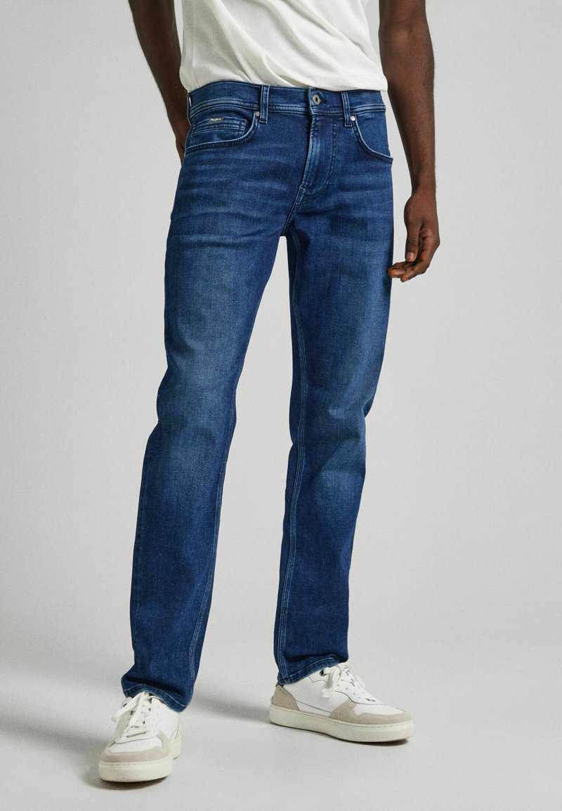 Jeans de mezclilla azul oscuro con corte recto, que cuentan con cinco bolsillos y un suave desgaste. Combinados con zapatillas blancas y una camiseta ligera.