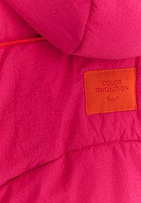 Růžová prošívaná bunda s oranžovou štítkem s vyraženým nápisem "COLOR REVOLUTION." Materiál působí lehce a má hladkou texturu.