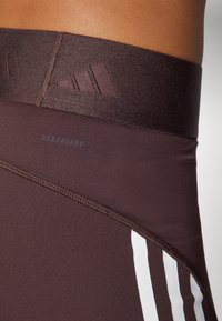 Bruna högmidjade träningsleggings med en strukturerad midja och tre vita ränder på sidan. Har "AEROREADY"-logotyp i blått.
