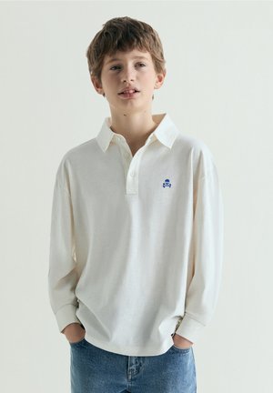 Jongen met kort bruin haar die een wit poloshirt met lange mouwen draagt met een klein blauw schedellogo en blauwe spijkerbroek, handen in de zakken.
