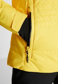 Gelbe gepolsterte Jacke mit einer seitlichen Reißverschlusstasche. Sie verfügt über ein schwarzes Innenfutter und eine glatte Stoffoberfläche. Goldfarbene Hardware-Akzente.