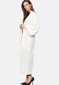 Lång vit stickad cardigan med ett strukturerat mönster, översize ärmar, sidofickor och en öppen design. Bärs över svarta kläder och klackar.