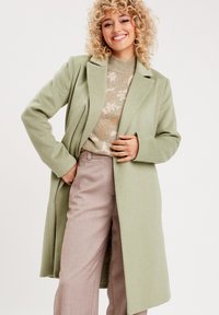 Manteau vert clair en mélange de laine avec un col à revers, des manches longues et une ouverture sur le devant, porté par-dessus un pull beige à motif et un pantalon clair.