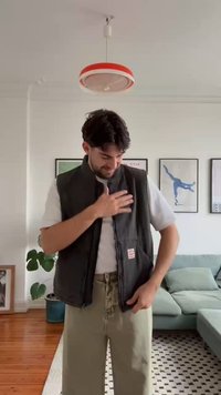 Siv prsluk bez rukava s prednjim zatvaračem i bočnim džepovima, nošen preko bele košulje. Kaki pantalone i šarena prostirka u svetloj sobi.