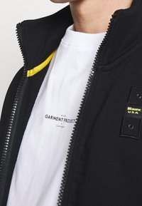 Svart zip-jacka med en gul accent vid kragen, bärs över en vit t-shirt med "Garment Project"-logotyp. Dragkedjedetalj synlig.