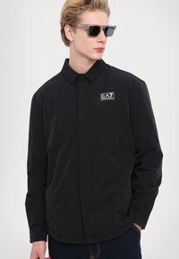 Svart lättviktjacka med kragdesign, med en logotyp på bröstet och slät textur. Modellen bär solglasögon.