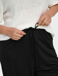 Černé kalhoty s nastavitelným stahovacím pasem, spárované s volným pleteným bílým topem. Zaměřte se na detail stahovací šňůrky.