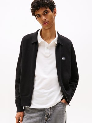 Zwarte ritska cardigan met geribde manchetten en zoom, gedragen over een witte poloshirt. Beschikt over een klein logo op de linkerborst en een gestructureerde stof.