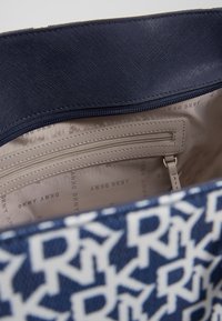 Kabelka DKNY s modro-bílým logovým vzorem, texturovaným tmavě modrým vnějškem a hladkým béžovým vnitřkem s kapsami na zip i volnými kapsami.