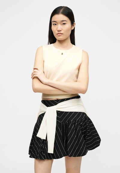 Νεαρή γυναίκα με μακριά σκούρα μαλλιά, που φοράει μπεζ τοπ χωρίς μανίκια, μαύρη φούστα με λεπτές ρίγες και ελαφρύ πουλόβερ δεμένο γύρω από τη μέση της.