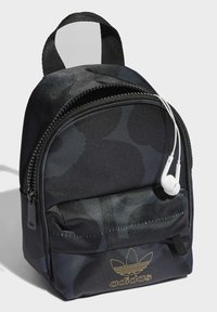 Black fabric backpack with a rounded shape, featuring a zippered compartment, front pocket, and gold Adidas logo. Includes white earphones attached.