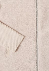 Vaaleanbeige fleece-takki, jossa on teksturoitu pinta. Varustettu ribbettyillä hihoilla ja hopeisella vetoketjuyksityiskohdalla edessä.