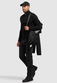 Svart stickad tröja med hög krage, svarta jeans och slitstarka svarta kängor. Håller en svart puffjacka och har på sig en svart keps.