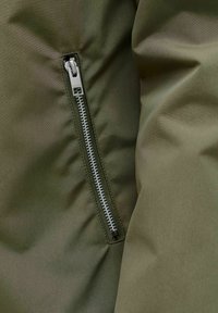 Oliverohelisest kergelt siledast kangast jakk; külgedel hõbedase tõmblukuga, millel on dekoratiivne tõmbetõmmits; lihtsa disainiga.