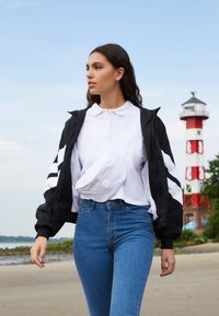 Jachetă neagră cu fermoar, cu dungi albe, cămașă polo albă, blugi albaștri cu talie înaltă și o geantă albă tip fanny pack. Cadru natural în aer liber.