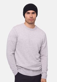 Pull léger gris clair avec des poignets et un ourlet côtelés, fabriqué en tissu doux. Bonnet en tricot noir sur la tête. Le mannequin se tient avec les mains dans les poches.