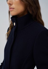 Manteau en laine bleu marine avec un col haut, une texture subtile et un détail en bouton-pression doré sur le devant.