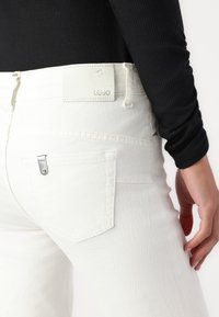 Jeans in denim bianco con una texture liscia, caratterizzati da un taschino posteriore con accessori in argento e un'etichetta Liu Jo sulla vita.