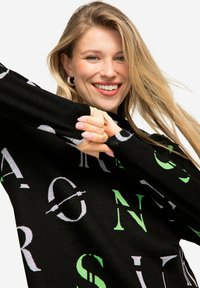 Schwarzer Pullover mit auffälliger, mehrfarbiger Schrift in Grün und Weiß. Das Material wirkt weich und strukturiert, mit lockerer Passform und langen Ärmeln.