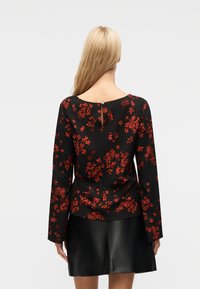 Femme aux longs cheveux blonds portant un haut à manches longues noir à fleurs rouges et une jupe noire, vue de dos.