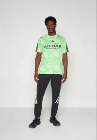 Žalios gėlėtos Adidas sporto marškinėliai su juodu ženklu, derinami su juodomis kelnėmis, turinčiomis baltas juosteles šoninėse.