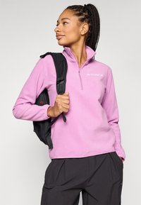 Lys rosa fleecegenser med kvart-glidelås og brodert logo; modell holder en svart ryggsekk og har på seg mørke bukser.