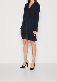 Navyblaues Kleid mit langen Ärmeln, einem V-Ausschnitt und einer Taillennaht. Das Kleid hat eine weiche Textur und wird mit schwarzen High-Heels kombiniert.