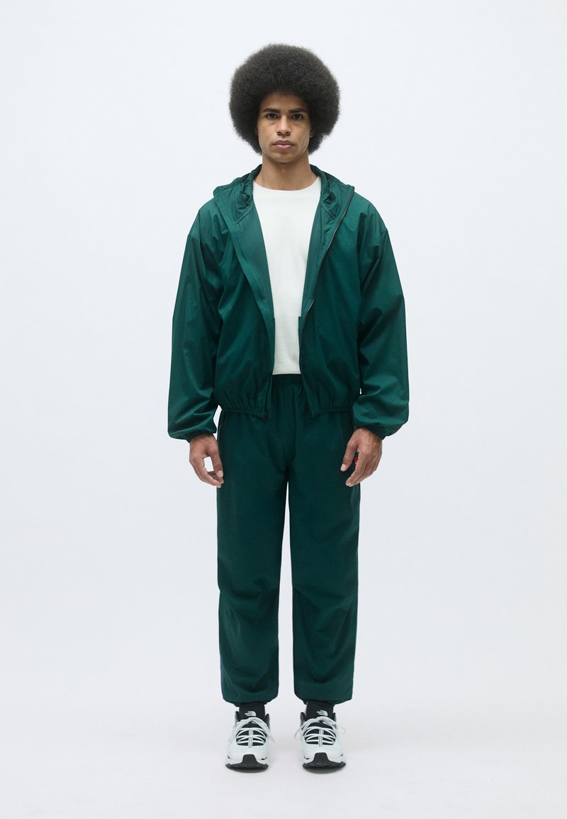 Grüner leichter Trainingsanzug mit kapuzenbehafteter, mit Reißverschluss versehenen Jacke und Hose mit elastischem Bund. Weißes Shirt darunter und schwarz-weiße Turnschuhe.