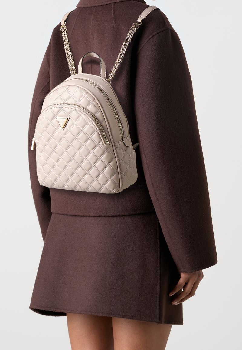 Personne portant un manteau marron et une jupe avec un petit sac à dos matelassé beige avec des bretelles chaîne dorée et un logo triangulaire au dos.
