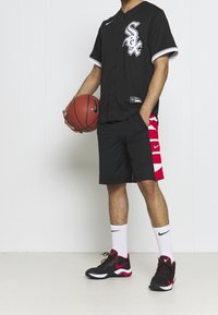 Maillot de sport noir avec des accents blancs et un logo, shorts noirs avec des motifs rouges sur le côté, chaussures de basket noires et rouges, tenant un ballon de basket.