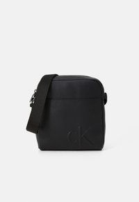 Bolso cruzado negro hecho de cuero liso, con cierre de cremallera, correa ajustable y logotipo en relieve en un lado. Diseño rectangular simple.