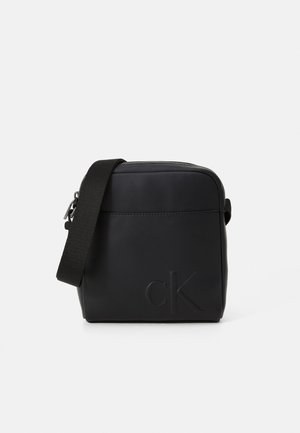 Sac bandoulière noir en cuir lisse, avec fermeture éclair, sangle ajustable et logo embossé sur un côté. Design rectangulaire simple.