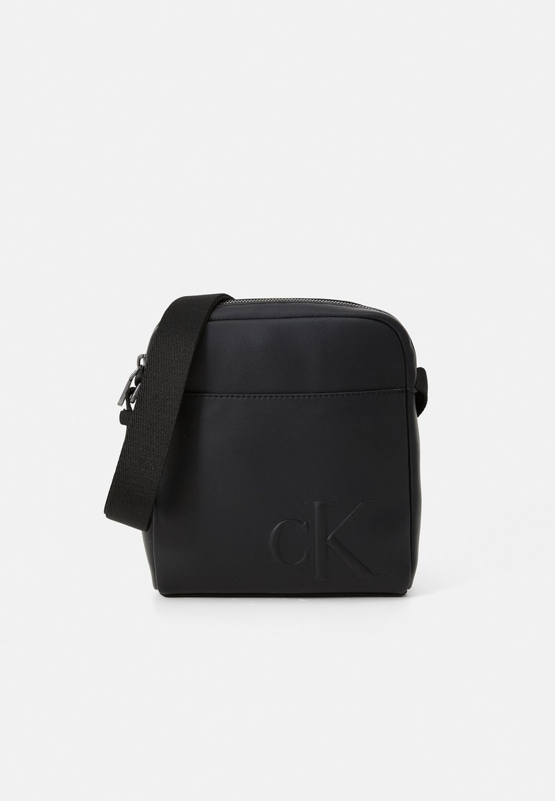 Bolso cruzado negro hecho de cuero liso, con cierre de cremallera, correa ajustable y logotipo en relieve en un lado. Diseño rectangular simple.