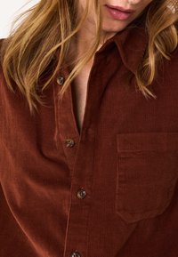 Camicia in velluto a coste marrone con colletto, chiusura a bottoni e una tasca sul petto, caratterizzata da un motivo a coste testurizzato.