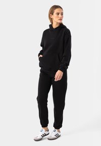 Melna kokvilnas kapuce ar priekšējo kabatu, apvienota ar atbilstošajām biksēm. Modelis valkā baltas kedas ar melniem un pelēkiem akcentiem. Vienkāršs, tīrs dizains.