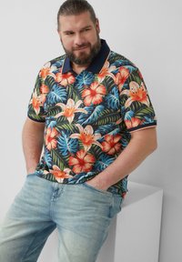 Korte mouw polo shirt met een levendig bloemenpatroon van oranje en witte lelies, donkergroene bladeren en een marineblauwe kraag. Lichtblauwe jeans.