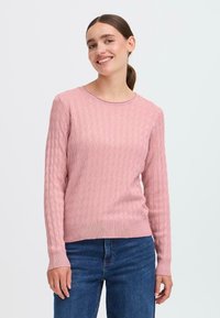 Lys rosa genser med teksturert kabelstrikkmønster, rund hals, ribbestrikkede mansjetter og nederkant, kombinert med blå denimjeans.