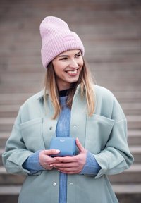 Mint green oversized jacket, light blue ribbed sweater, pink knit beanie. Holding a small blue wallet with a logo, standing against gray steps.