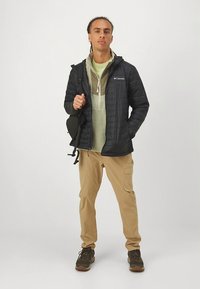 Must kapuutsiga tikitud jakk, helerohelise kihi ja topp, khaki värvi võrgutrikood ning pruunid matkajalatsid. Inimene hoiab käes väikest musta kotti.