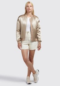 Béžová saténová bomber bunda s ribkovanými manžetami, nošená přes bílý top a krémové šortky, doplněná béžovými teniskami. Hladká textura, ležérní design.