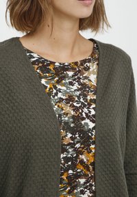 Grön texturerad kofta över en mönstrad topp med jordnära färger och blommönster som framhäver kontrasten mellan material och färger.