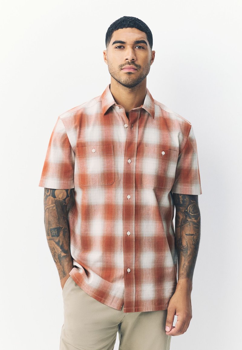 Camicia a maniche corte con bottoni, a quadri arancioni. Presenta due tasche frontali e una texture liscia con quadri bianchi e rossi.