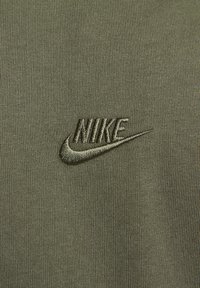 Nike logo izšūts tumši zaļā diegā uz olīvu zaļas teksturētas auduma fona.