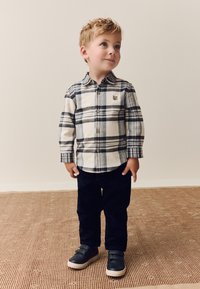 Flanellhemd in Creme und Marinekaromuster, geknöpft, mit einem kleinen Bären-Emblem; kombiniert mit schwarzen Cordhosen und schwarzen Turnschuhen mit beigen Sohlen.