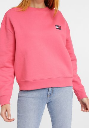 Kobieta ubrana w jasnoróżową bluzę Tommy Jeans z prążkowanymi mankietami i ściągaczem, w połączeniu z jasnoniebieskimi jeansami.